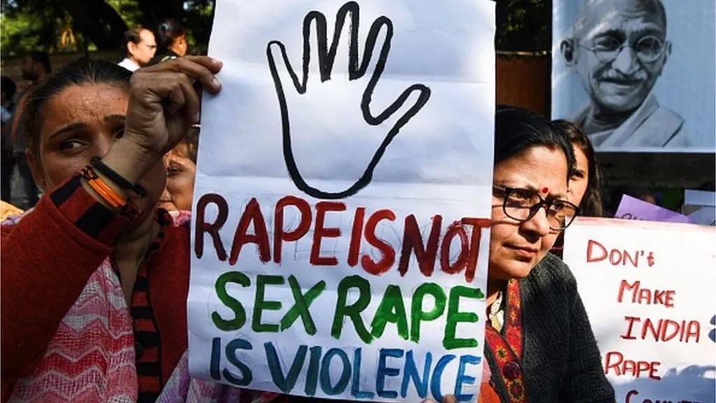 Two feminine-presenting individuals in the middle of a protest demonstration holding a placard saying "RAPE IS NOT SEX RAPE IS VIOLENCE".