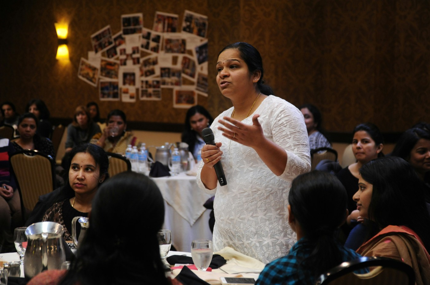 Several women sit around tables at a venue, with one of them stood and talking into the mic.