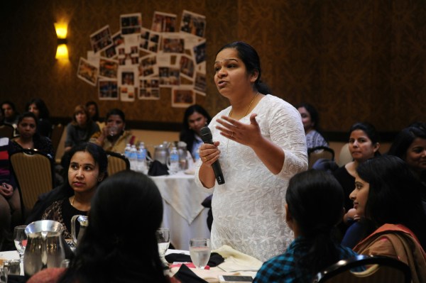 Several women sit around tables at a venue, with one of them stood and talking into the mic.