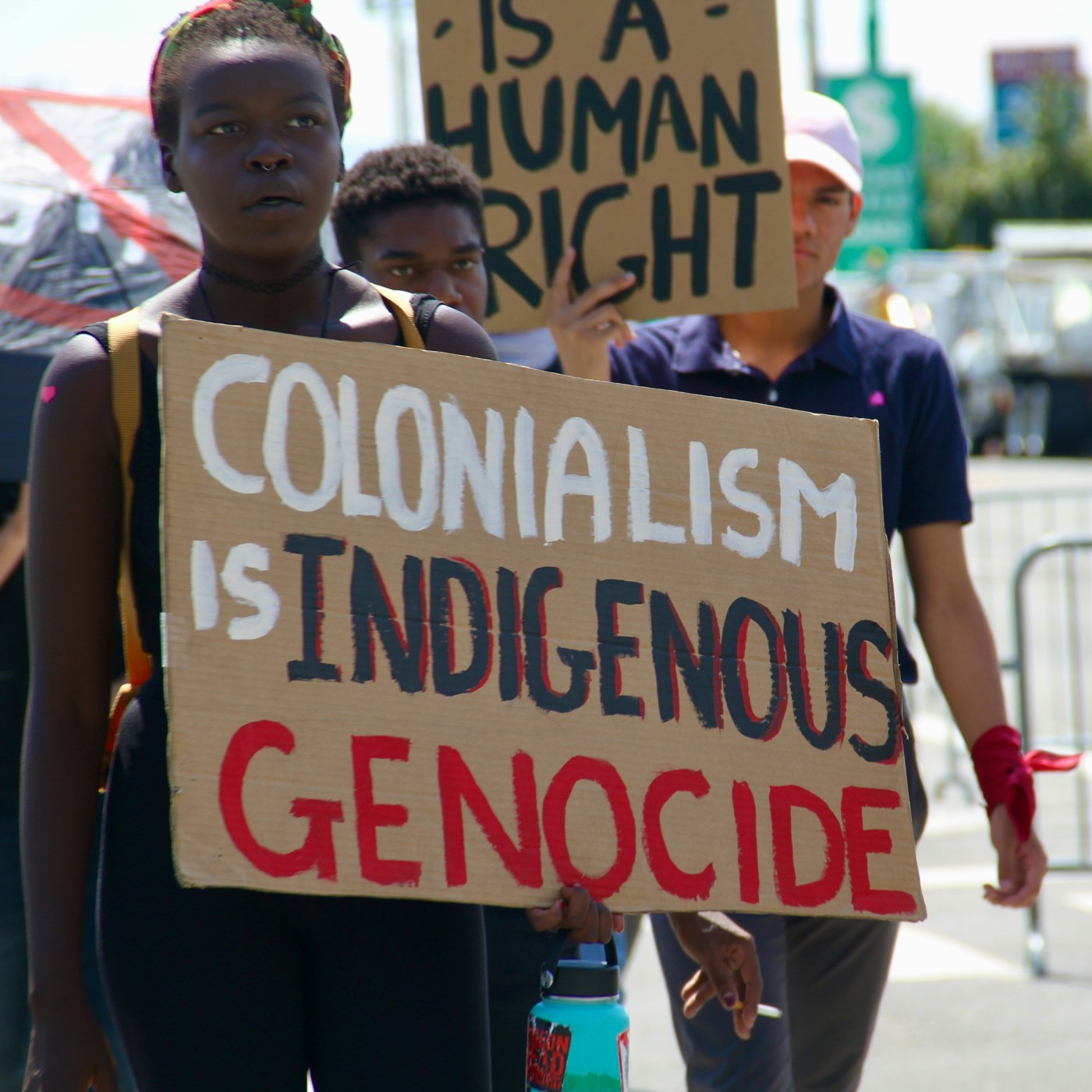 Several people at a protest. One in the front holds a placard saying "Colonialism is Indigenous Genocide".